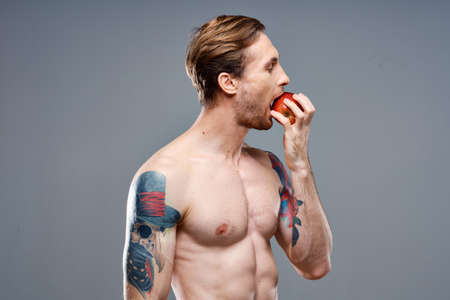 sexy athlete with pumped up muscles eats an apple on a gray background and a tattoo