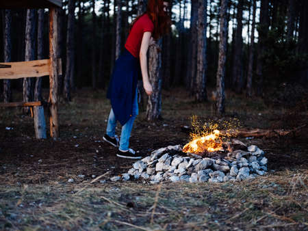 A romantic woman in jeans and a T-shirt sits near a campfire in natureの写真素材