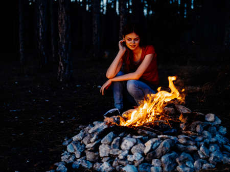 A woman sits near the fire and looks at the fire in a pine forestの写真素材