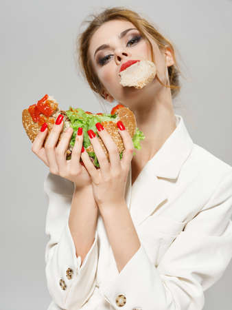 woman with red manicure eating hamburger and makeup on face light jacket on gray background