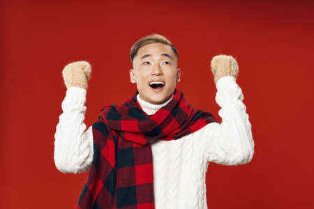happy young man in a sweater and scarves on a red background holds his hands in a fist
