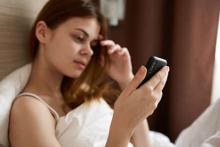 portrait of a woman in bed under a white blanket with a mobile phone and design interiorの写真素材