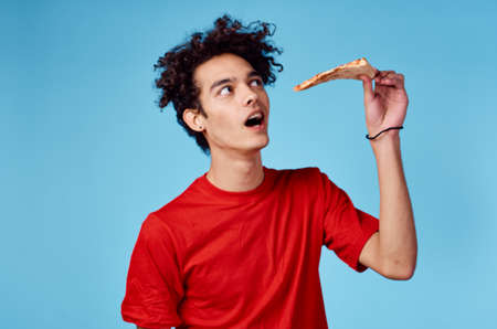 man with a piece of pizza in his hand on a blue background teenager in a red t-shirt curly hairの写真素材