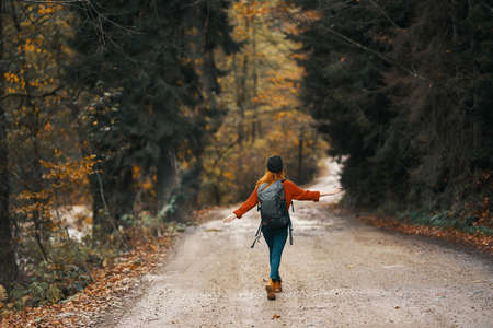 woman in jeans jacket with a backpack walks along the road in the autumn forest
