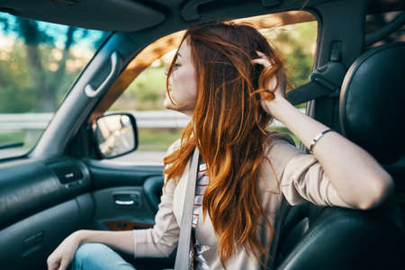 happy red-haired woman in a beige coat in the front seat of a car smile model gesturing with her...