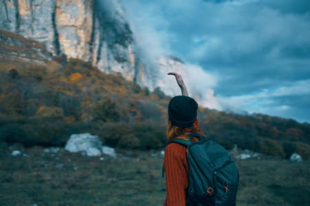 a traveler with a backpack and in a red sweater shows the mountains with her hand and the blue sky cloudsの写真素材