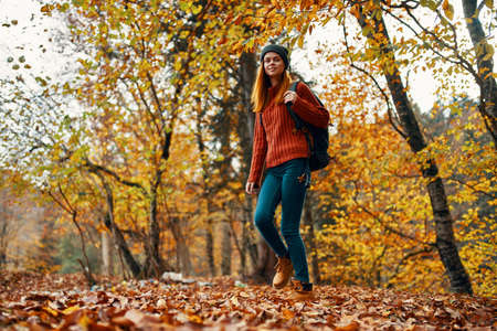 young woman in jeans and a sweater with a backpack on her back walks in the park in autumn in...