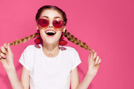 cheerful woman in sunglasses holding pigtails pink background fashionable clothes summerの写真素材