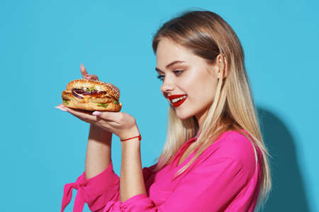 cheerful blonde in a pink shirt hamburger fast food snack blue backgroundの写真素材