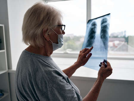 an elderly woman in a medical mask and glasses examines an x-ray near the window of a covid-19 passportの写真素材