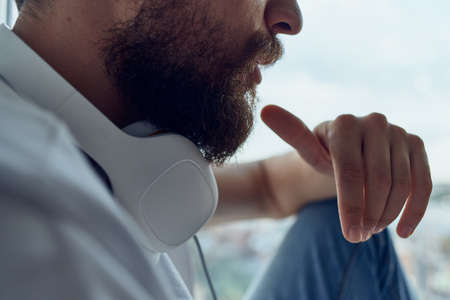 man in white t-shirt sitting near the window in headphones listening to music