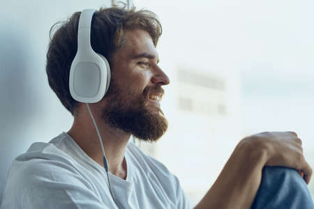 man in white t-shirt sitting near the window in headphones listening to musicの写真素材