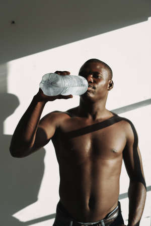 African man with a pumped-up torso drinks water from a bottle on a light background roomの写真素材