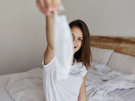 happy woman with medical mask in hand sitting on bed in a light roomの写真素材