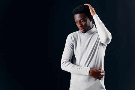a man of African appearance in a gray sweater holds his head against a dark backgroundの写真素材