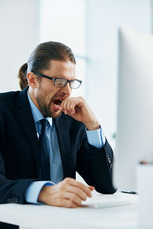 business man in a suit sitting at his desk tired in front of a computerの写真素材