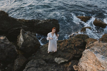 A woman in a white dress stands barefoot on the stones on the ocean shore top viewの写真素材