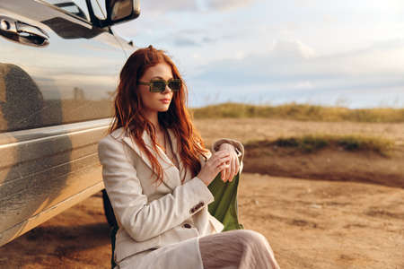 beautiful woman sitting on a chair in a field wearing dark glasses near the car endless fieldの写真素材