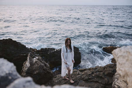 A woman in a white dress stands on rocky stones by the oceanの写真素材
