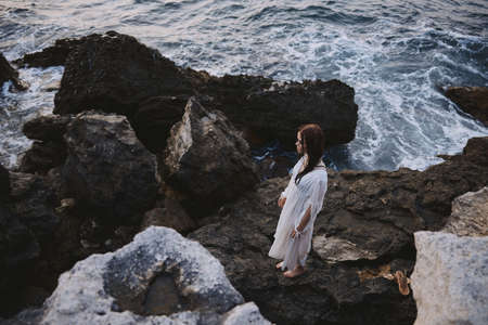 A woman in a white dress with wet hair stands on the stones by the ocean top viewの写真素材