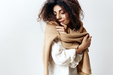 Curly Pretty Latin Female in White Shirt Posing Isolated Hugging Herself Enjoying Warm Sweater Closing Eyes At White Studio Wall Background. Fashion Seasonal Sale New Collection Concept. Copy spaceの写真素材