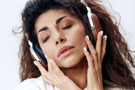 Enjoyed Curly Pretty Latin Woman in White Shirt In Headphones Closing Eyes Isolated At White Studio...