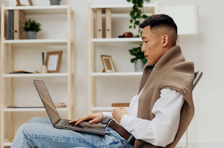 Asian man sitting in a chair in a room with a laptop on his lap Gray background