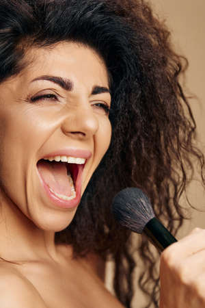 Overjoyed pretty adorable tanned sexy curly Latin makeup artist hold brush like micro singing relaxing satisfied posing isolated on pastel beige background close eyes. Cosmetic offer concept closeupの写真素材