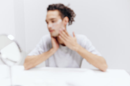 handsome guy in a white T-shirt sits at a table with a mirror and a towel cream on his face skin healthの写真素材