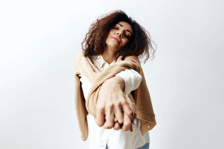 Curly Pretty Latin Female in Pastel Warm Sweater and White Shirt Raising Crossed Hands At Camera...