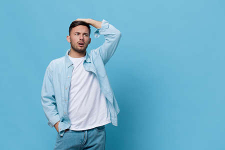 Worried sad unhappy tanned handsome man in casual basic t-shirt touch head posing isolated on blue studio background. Copy space Banner Mockup. People emotions Lifestyle concept. Lucky winnerの写真素材