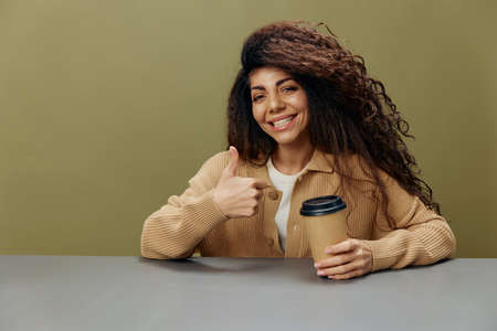 Cheerful smiling tanned curly Latin female hold for a cup to-go gesture like sit at the table...