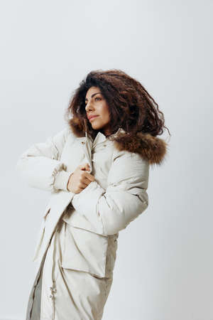 Serious Curly Pretty Latin Lady Wrapping In Soft Warm Beige Winter Coat With Fur Hood Posing Isolated Looking Aside At White Studio Background. Fashion Huge Sale New Collection Concept Vertical Shotの写真素材