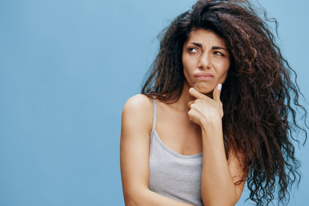 GREAT OFFER. Thoughtful beautiful Latin female with afro with hand near cheek, look at side. Fashion. Studio shoot over blue sky background. Copy space, free place for design adの写真素材