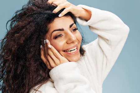 Happy Latin lady in bathrobe touch natural curly hair and clean flawless skin posing isolated on pastel blue background, feeling excited of effective beauty procedure. Closeup portrait, studio shotの写真素材