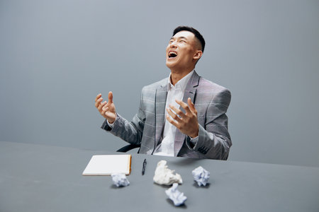 Asian man in a suit sits at a table emotions crumpled paper isolated background
