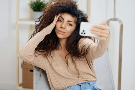 Cheerful awesome tanned curly Latin lady taking selfie touch head with trendy smartphone sit on...