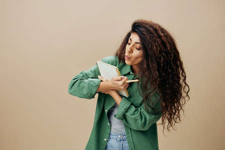 WRITER CONCEPT. Lovely tanned curly Latin female in love in casual things kiss notebook posing...