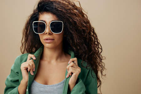 FASHION CONCEPT. Sexy adorable tanned curly Latin female in casual things sunglasses touch collar posing isolated over pastel beige background look at camera. Copy space Mockup Banner. Fashion offerの写真素材