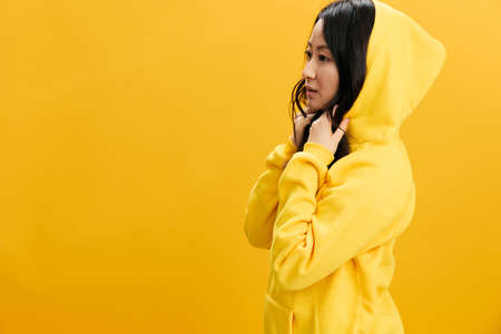 Adorable cute happy Asian student young woman in yellow sweatshirt puts on the hood posing isolated on over orange studio background. Cool offer. People Emotions concept. Copy space for adの写真素材