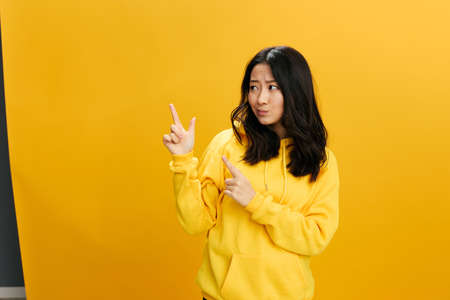 Pensive doubting Asian student young woman in yellow hoodie sweatshirt show at empty copy space for ad posing isolated on over orange studio background. Cool offer. People Emotions conceptの写真素材