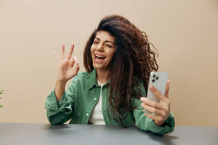 Joyful smiling happy tanned curly Latin lady show gesture Okay hold phone sit at the table isolated over pastel beige background winks at camera. Copy space Mockup Banner. Concept Mobile Appの写真素材