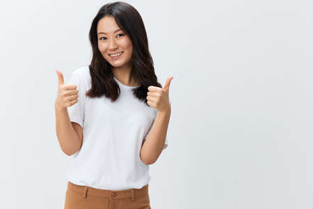 Cheerful happy cute Asian young female in white basic t-shirt show thumb up Like gesture both hand posing isolated on over white background. The best offer for ad. People Lifestyle concept. Free placeの写真素材