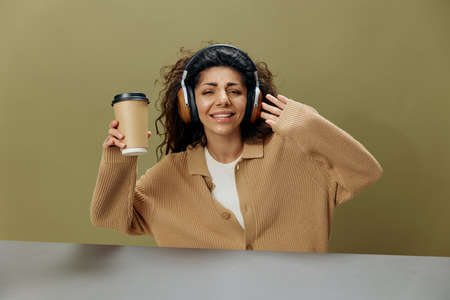 COOL MOOD CONCEPT. Enjoyed tanned curly Latin female in headphones with coffee cup to-go dance sing...