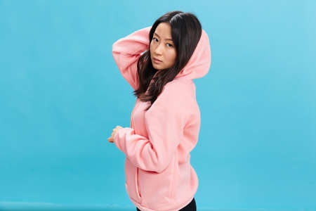 Side of view cute Asian student young lady in pink hoodie sweatshirt posing isolated on over blue...