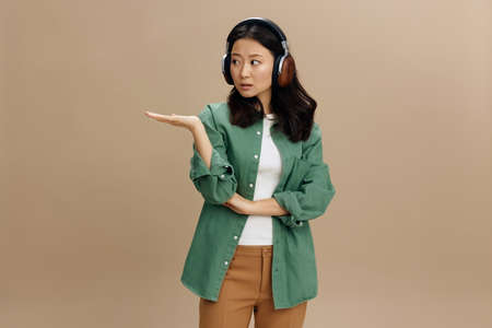 Wow, look here. Korean shocked young woman in khaki green shirt headphones hold palm up aside posing isolated on beige pastel studio background. Cool fashion offer. Music App Platform Ad conceptの写真素材