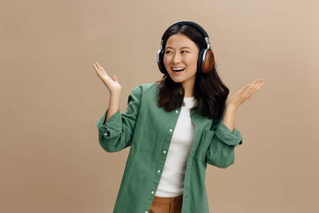 Hey look here. Cheerful happy Asian young woman in khaki green shirt headphones raise palms up posing isolated on over beige pastel studio background. Cool fashion offer. Music App Platform Ad conceptの写真素材