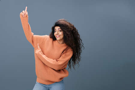 LOOK AT THIS CONCEPT. Enjoyed happy tanned curly Latin female in warm sweater show point fingers up...