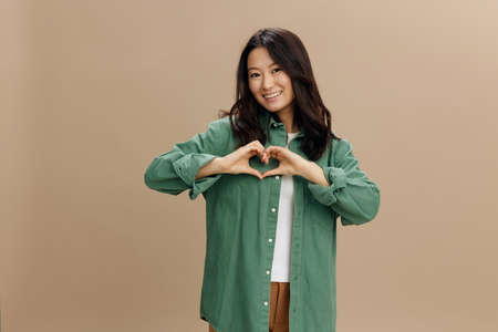 We all need Love. Cute happy Asian student young woman in khaki green shirt show heart shaped sign posing isolated on beige pastel studio background. Cool fashion offer. Lifestyle and Emotions conceptの写真素材