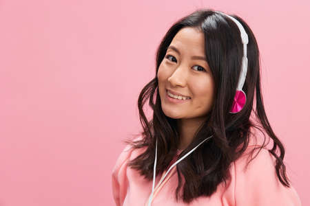 Smiling cutie cute Asian young lady in pink hoodie sweatshirt with cute headphones looks at you with love posing isolated on pink studio background. Good offer. Sound streaming platform ad conceptの写真素材